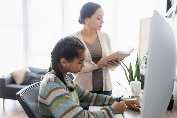 Africano americano ragazza scrittura in notebook vicino sfocato monitor e madre lettura libro — Foto stock