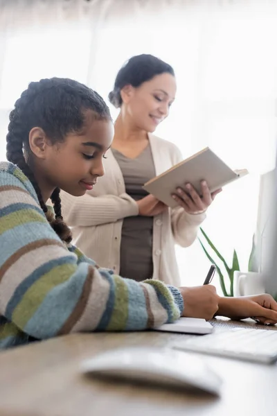 African american girl writing in notebook near mom reading book and computer mouse on blurred foreground — Stock Photo