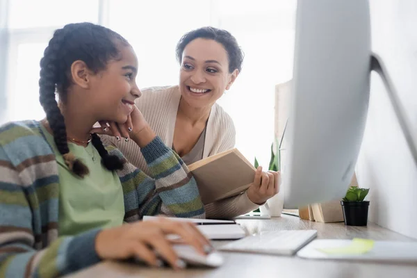 Glücklich afrikanisch-amerikanische Frau mit Buch sieht Tochter bei Hausaufgaben in der Nähe verschwommener Computer — Stockfoto