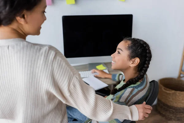 Happy african american girl looking at blurred mother while doing homework near monitor with blank screen — Stock Photo