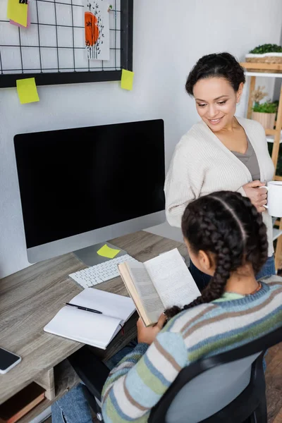 Africano americano menina leitura livro perto monitor com tela em branco e sorrindo mãe com xícara de chá — Fotografia de Stock