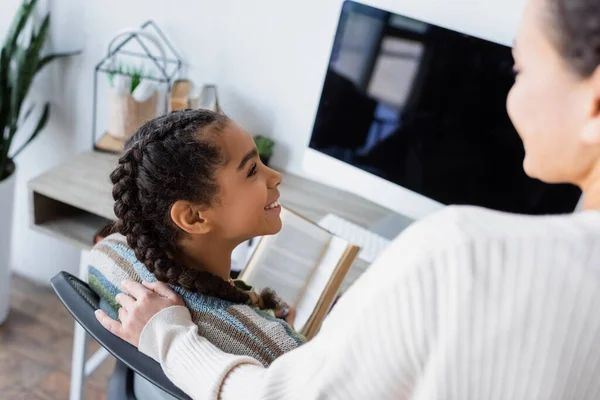 Femme afro-américaine floue debout près de fille souriante assis avec livre près du moniteur avec écran blanc — Photo de stock