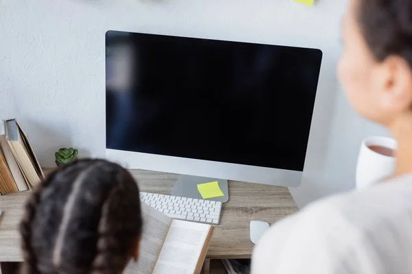 High angle view of african american girl reading book near monitor with blank screen and blurred mother — Stock Photo