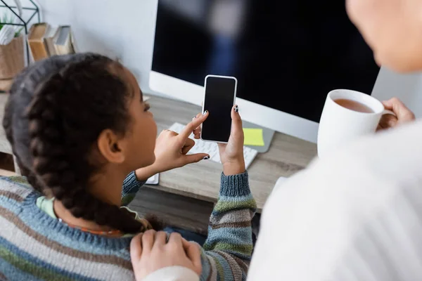 Afro-américaine fille à l'aide d'un téléphone portable avec écran vide près de l'ordinateur moniteur et maman floue avec tasse de thé — Photo de stock