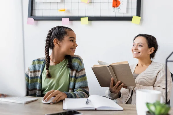 African american teenage girl smiling near computer and blurred mother with book — Stock Photo