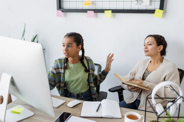 Afrikanerin gestikuliert, während sie mit Buch auf Computermonitor neben Mutter schaut — Stockfoto