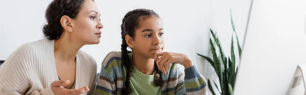 Mujer afroamericana apuntando a monitor de computadora cerca de hija reflexiva, pancarta - foto de stock