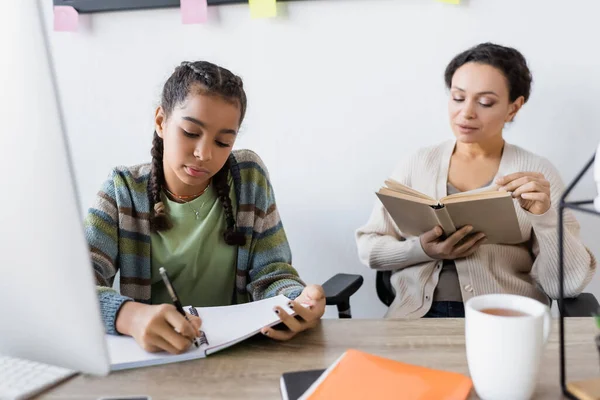 Afrikanisch-amerikanisches Teenager-Mädchen schreibt in Notizbuch in der Nähe verschwommener Computer und Mutter liest Buch — Stockfoto