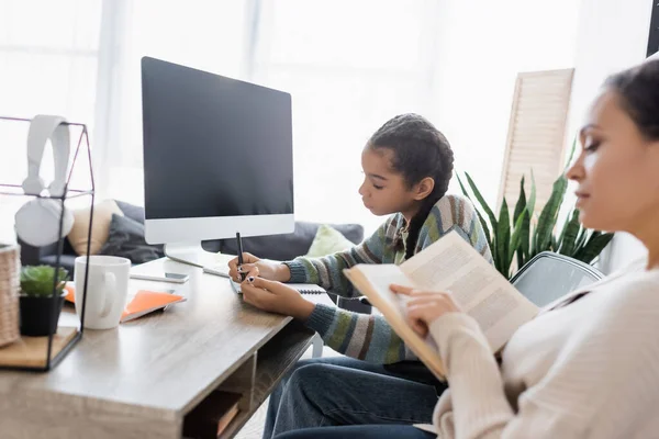 Afro-américaine livre de lecture femme tandis que la fille écrit dans un ordinateur portable près de moniteur avec écran blanc — Photo de stock