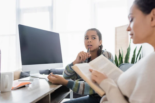 Blurred african american woman reading book near thoughtful daughter and monitor with blank screen — Stock Photo