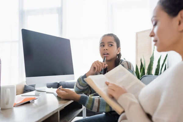 Borrosa mujer afroamericana leyendo libro cerca de hija reflexiva y monitor con pantalla en blanco - foto de stock