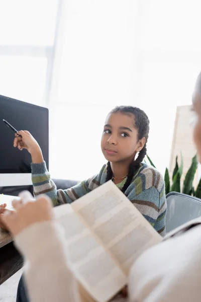 Afroamericana adolescente apuntando con pluma en monitor con pantalla en blanco cerca borrosa mamá con libro - foto de stock