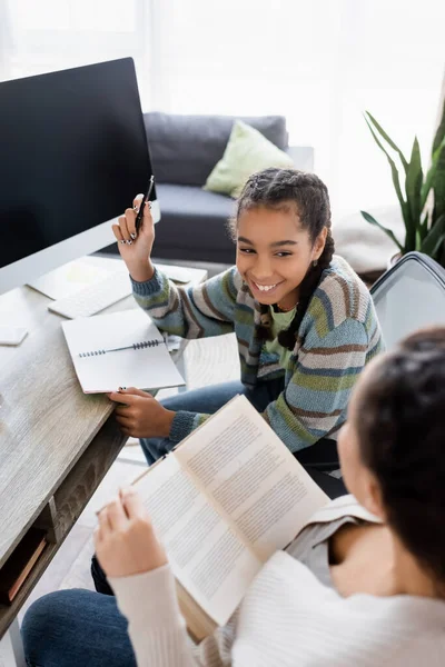 Усміхнена афроамериканська дівчина вказує ручкою на монітор з порожнім екраном біля розмитої мами з книгою — стокове фото