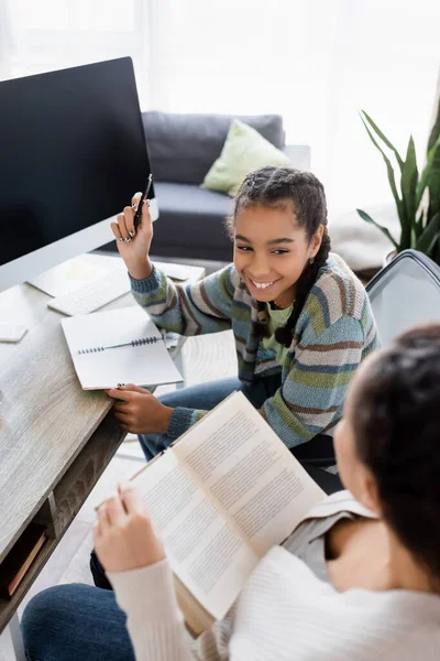 Lächelnde Afroamerikanerin zeigt mit Stift auf Monitor mit leerem Bildschirm nahe verschwommener Mutter mit Buch — Stockfoto