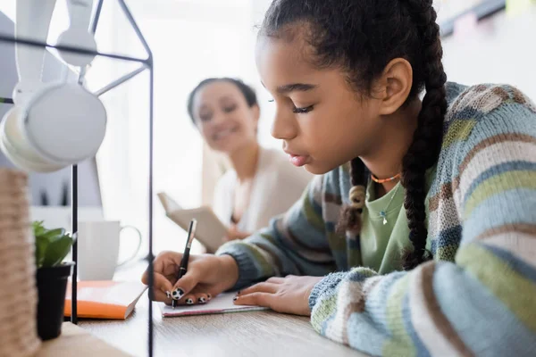 Chica afroamericana escribiendo en un cuaderno mientras hace la tarea cerca de los auriculares y mamá borrosa - foto de stock