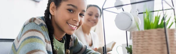 Glücklich afrikanisch-amerikanisches Teenager-Mädchen, das in die Kamera schaut, während es Hausaufgaben in der Nähe der verschwommenen Mutter macht, Banner — Stockfoto