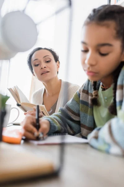 Offuscata ragazza adolescente africana americana che scrive in notebook vicino alla madre con libro — Foto stock