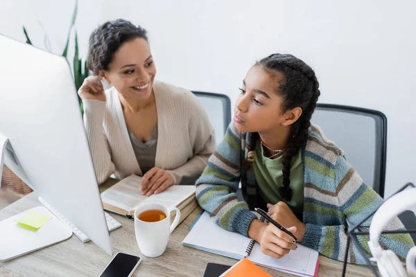 Lächelnde Afroamerikanerin hilft Teenager-Tochter bei Hausaufgaben in der Nähe des Computermonitors — Stockfoto