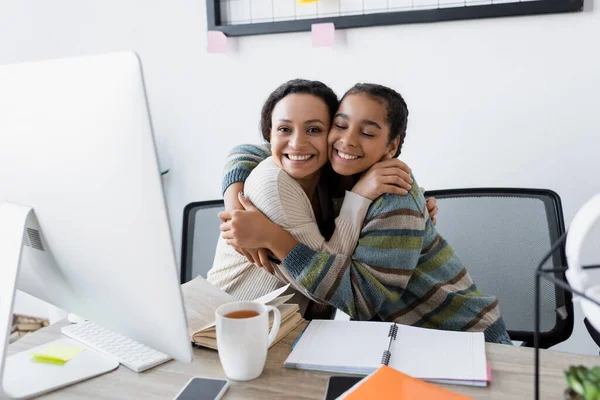 Glückliche afrikanisch-amerikanische Mutter und Tochter umarmen sich in der Nähe von Computer und Notebooks — Stockfoto