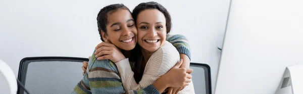 Mujer afroamericana feliz y adolescente abrazando cerca del monitor de la computadora, bandera - foto de stock