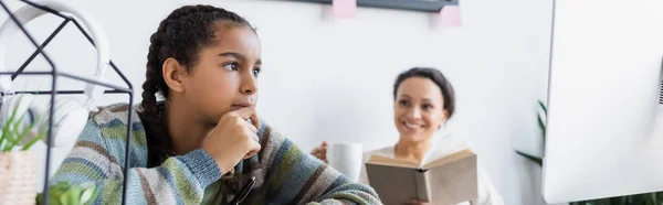 African american teen girl thinking near computer monitor and smiling mother with book on blurred background, banner — Stock Photo