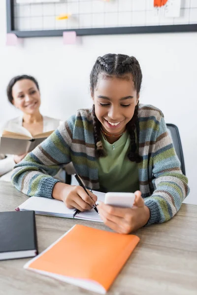 Glücklich afrikanisch-amerikanisches Teenager-Mädchen schreibt in Notizbuch nahe Mutter mit Buch lächelnd auf verschwommenem Hintergrund — Stockfoto