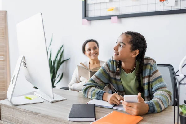 Sorridente ragazza africana americana che scrive nel taccuino vicino al monitor del computer e mamma offuscata con il libro — Foto stock