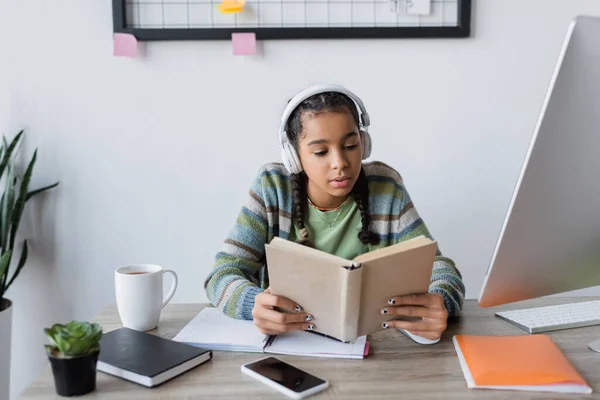 Africano americano adolescente in cuffie lettura libro vicino al computer e smartphone con schermo bianco — Foto stock