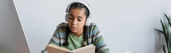 African american teenage girl in headphones reading book while studying at home, banner — Stock Photo