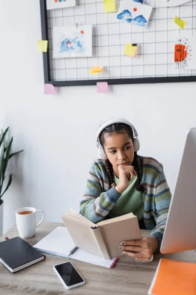 Nachdenkliches afrikanisch-amerikanisches Mädchen mit Kopfhörer, das Buch hält, während es auf den Monitor schaut — Stockfoto