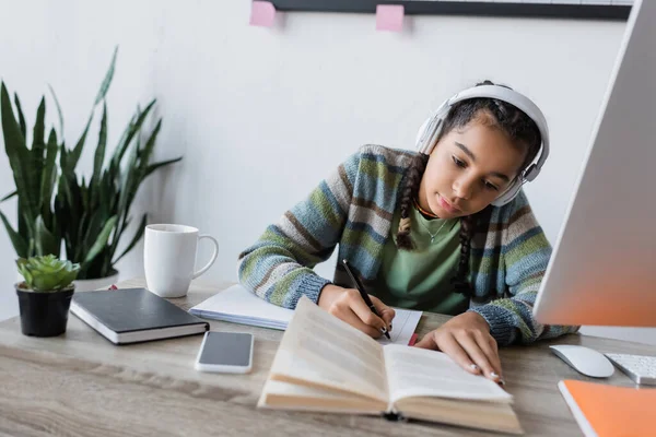 Menina afro-americana em fones de ouvido escrevendo perto de livro e computador enquanto estudava em casa — Fotografia de Stock
