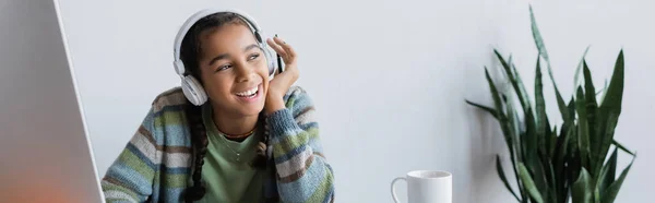 Щаслива афроамериканська дівчина в навушниках посміхається біля монітора комп'ютера вдома, банер — Stock Photo