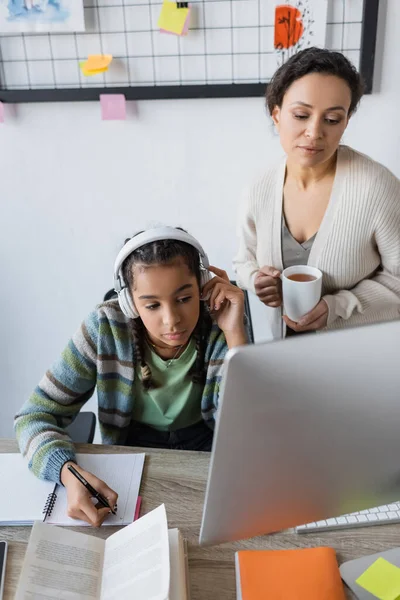 Chica afroamericana en auriculares escribir en portátil cerca de la computadora y la madre con taza de té - foto de stock
