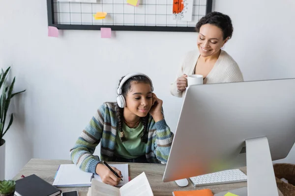 Menina americana africana em fones de ouvido escrevendo em notebook perto do monitor e sorrindo mãe com xícara de chá — Fotografia de Stock