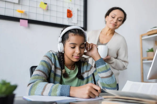 African american girl in headphones writing in notebook near blurred mom with cup of tea — Stock Photo