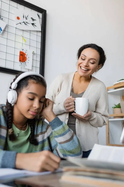 Afrikanisch-amerikanisches Mädchen mit Kopfhörern bei Hausaufgaben neben lächelnder Mutter mit einer Tasse Tee — Stockfoto