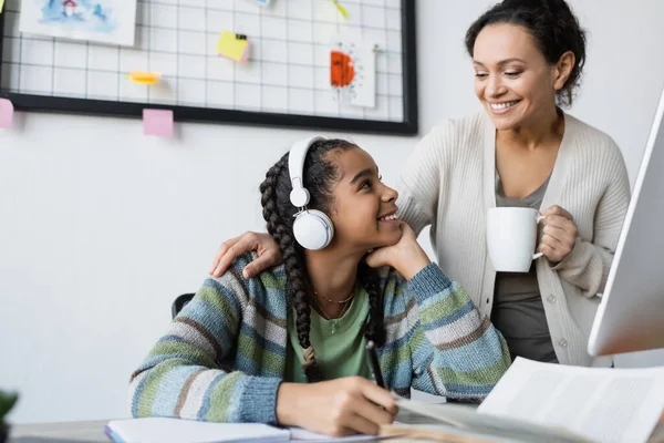 Africano americano chica escuchar lección en auriculares cerca sonriente mamá con taza de té - foto de stock