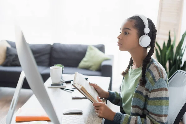 Afroamericana adolescente en auriculares mirando monitor de computadora mientras sostiene libro - foto de stock