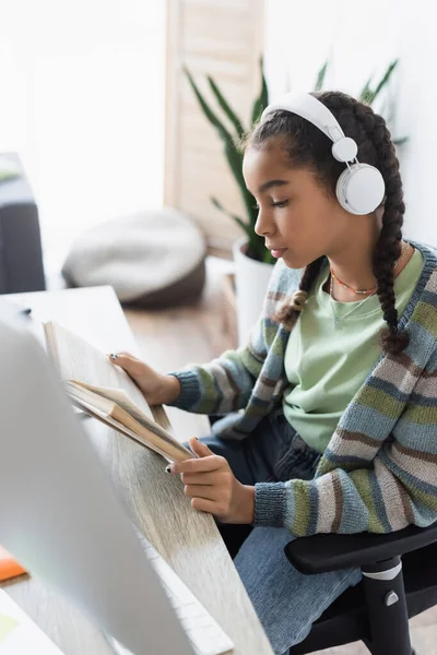 Africano americano estudante em fones de ouvido leitura livro perto de monitor de computador borrado — Fotografia de Stock