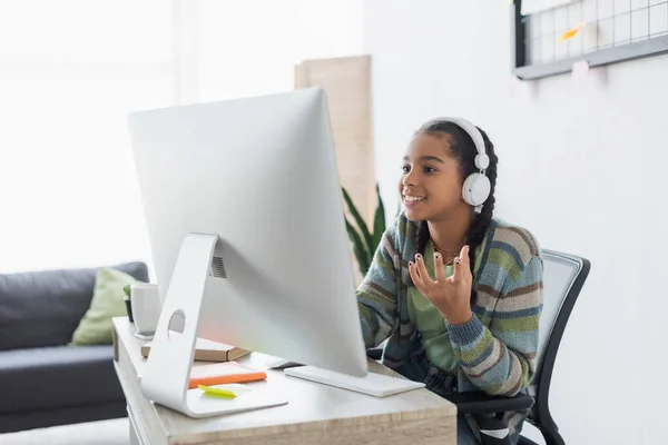 Fröhliches afrikanisch-amerikanisches Mädchen mit Kopfhörern gestikuliert in der Nähe des Computers zu Hause — Stockfoto