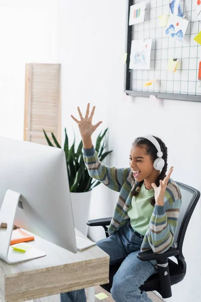 Eccitato africano americano giovanissima ragazza in cuffie mostrando wow gesto vicino computer monitor — Foto stock