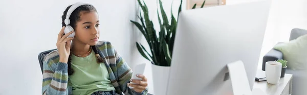 Menina americana africana concentrada em fones de ouvido segurando smartphone enquanto olha para o monitor, banner — Fotografia de Stock