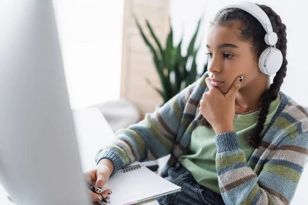 Reflexiva colegiala afroamericana en la escritura de auriculares en portátil cerca del monitor borroso de la computadora - foto de stock