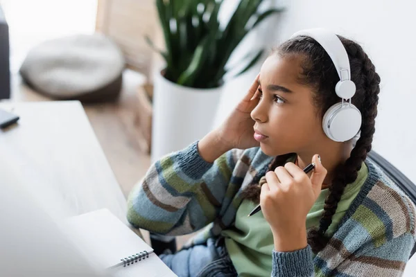 Pensativo afro-americano adolescente menina em fones de ouvido segurando caneta e olhando para longe enquanto estudava em casa — Fotografia de Stock