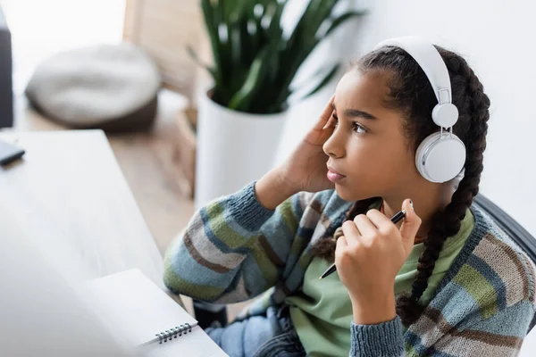 Réfléchi afro-américain adolescent fille en casque tenant stylo et détournant les yeux tout en étudiant à la maison — Photo de stock