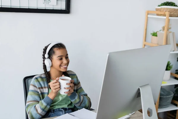 Menina americana africana feliz segurando xícara de chá enquanto sentado perto do monitor do computador em fones de ouvido — Stock Photo