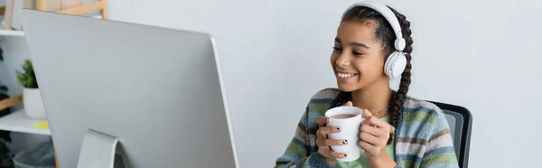 Joyful african american schoolgirl in headphones holding cup of tea near computer monitor, banner — Stock Photo