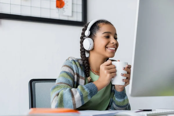 Happy african american schoolgirl in headphones holding tea cup while looking at monitor — Stock Photo