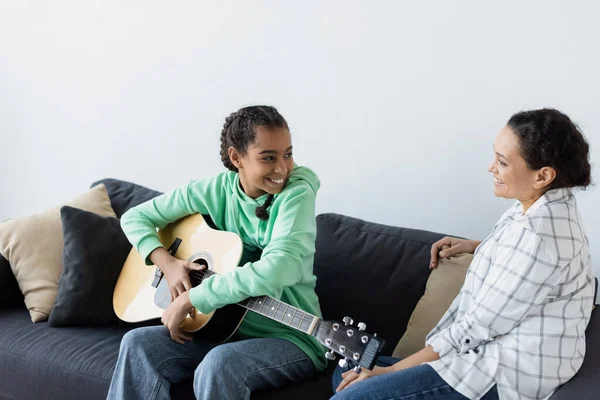 Счастливая африканская американская девочка-подросток с акустической гитарой, сидящей на диване рядом с улыбающейся мамой — стоковое фото