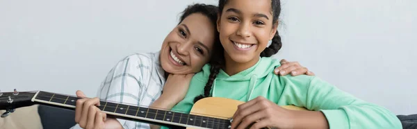 Freudige afrikanisch-amerikanische Frau umarmt Teenager-Tochter, die Gitarre spielt, Banner — Stockfoto